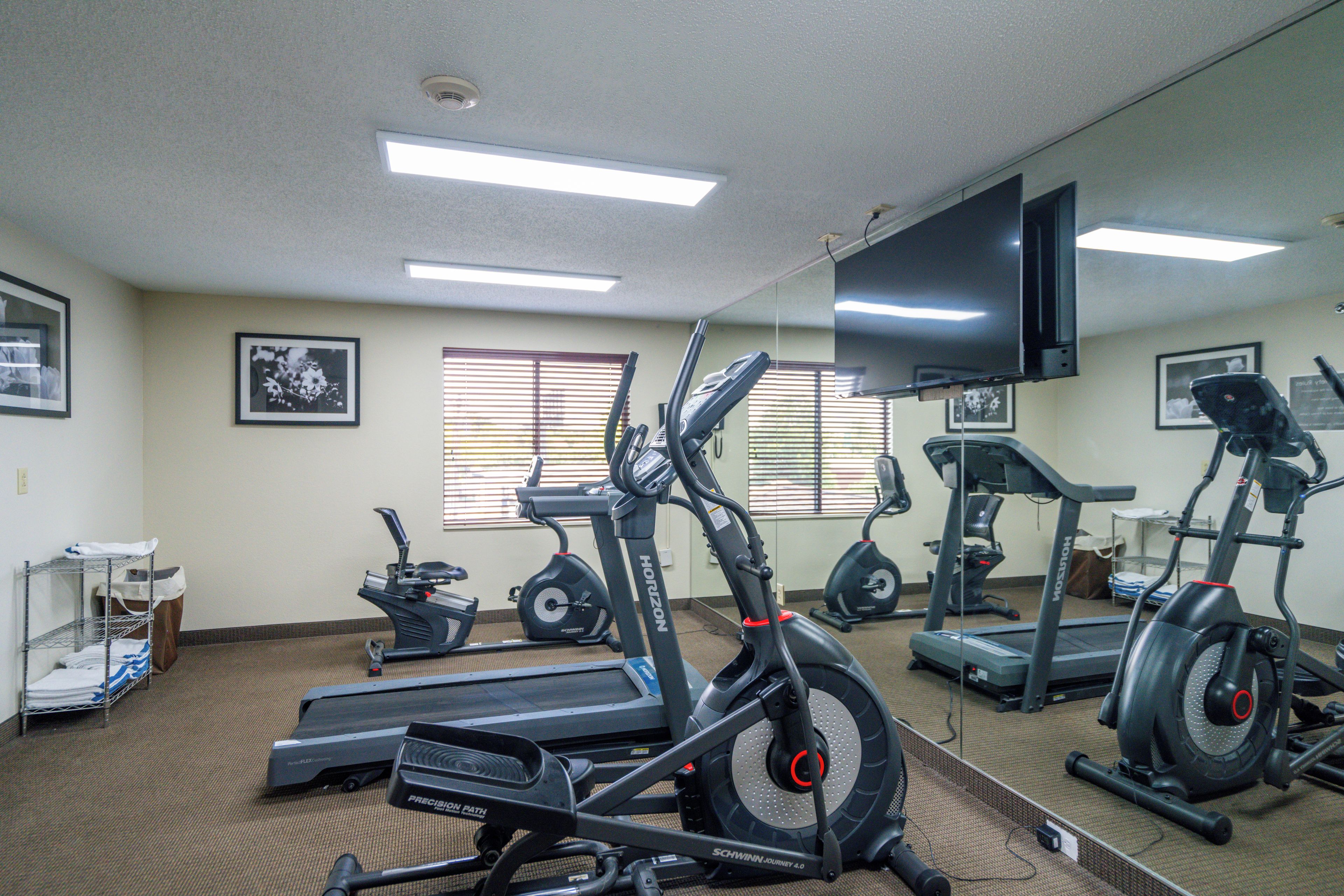 Un gimnasio con varias máquinas de ejercicio, un televisor en la pared y una ventana con persianas.