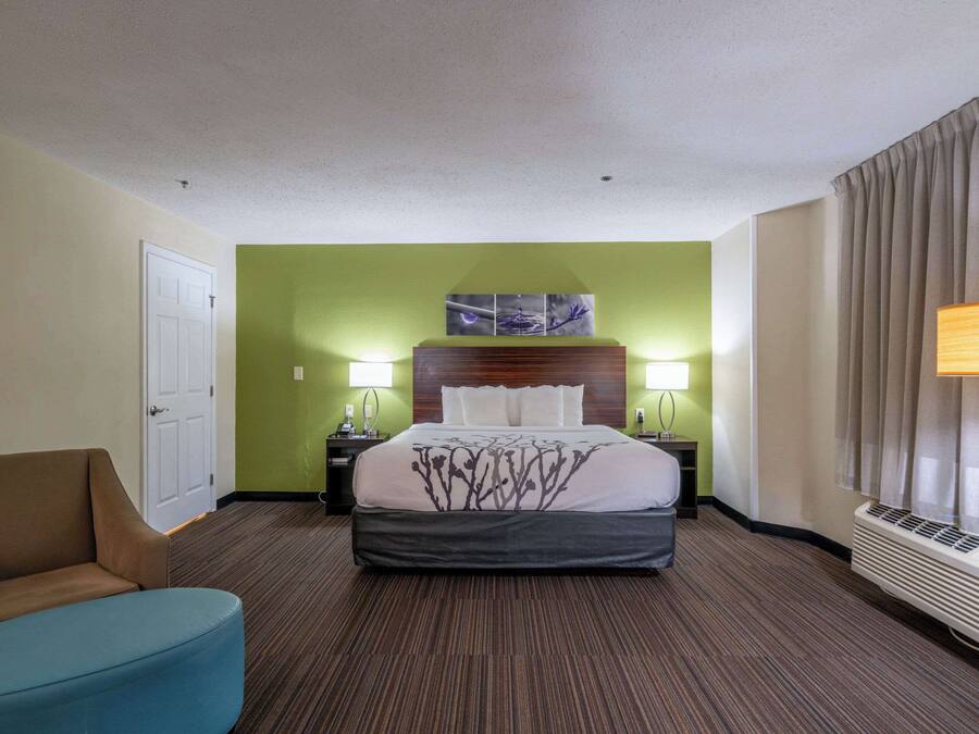 Une chambre d’hôtel avec un lit, une table de chevet, une lampe, une chaise et un mur orné d’un tableau.