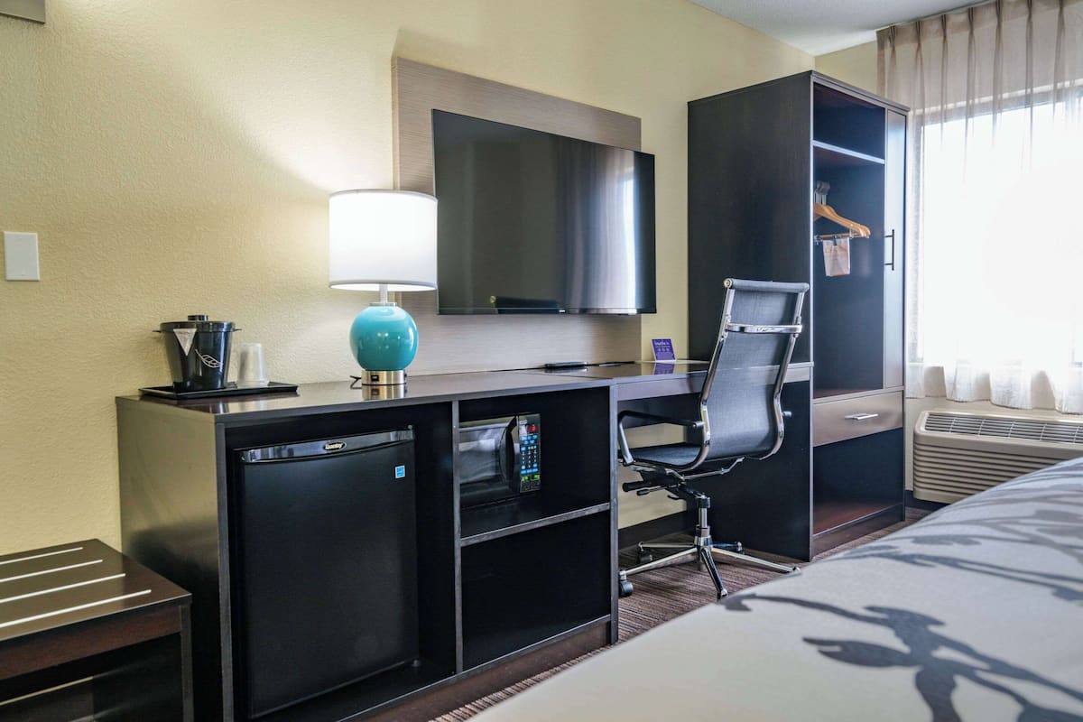 A hotel room with a desk, a chair, a TV, a mini-fridge, a lamp, and a wardrobe.