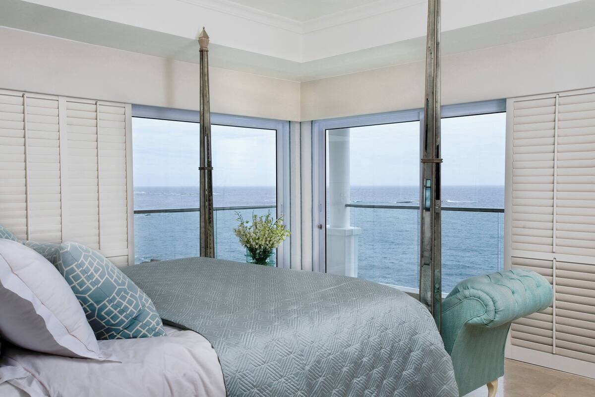 A bedroom with a large bed, a view of the sea through floor-to-ceiling windows, and a balcony with white shutters.