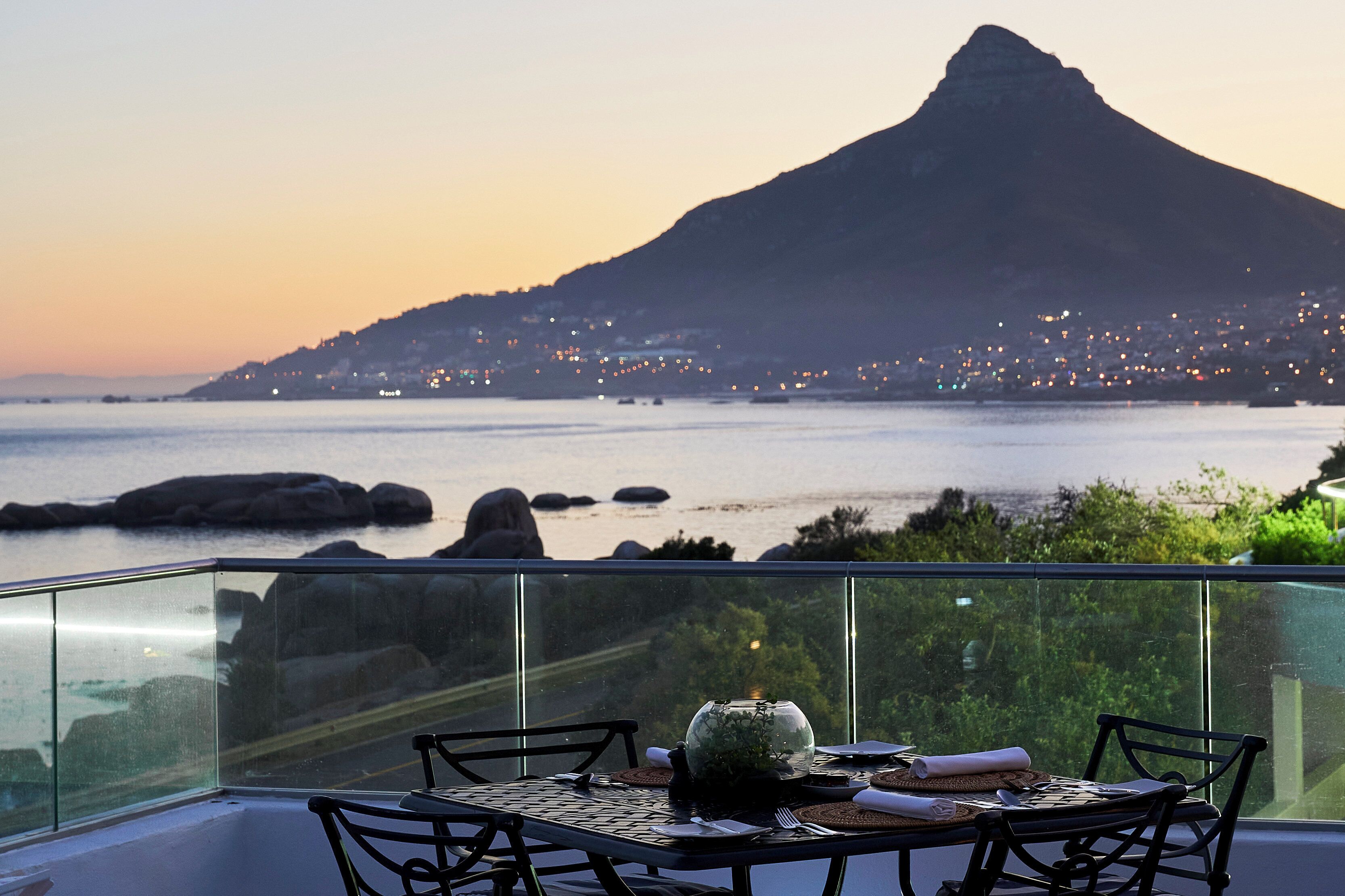 A dining table with chairs and a vase on a balcony overlooking a coastal cityscape with a prominent mountain in the background.