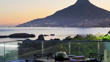 A dining table with chairs and a vase on a balcony overlooking a coastal cityscape with a prominent mountain in the background.