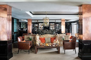 A luxurious lobby with a high ceiling, a central seating area with a patterned sofa, leather armchairs, and a coffee table with a tea service.
