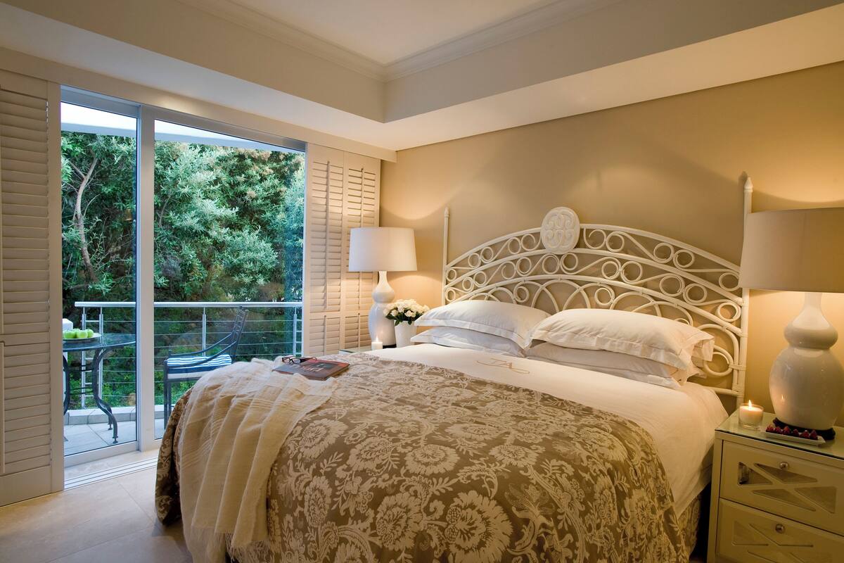 A bedroom with a large bed, a nightstand with a lamp, a glass table with a chair, and a balcony with a view of trees.