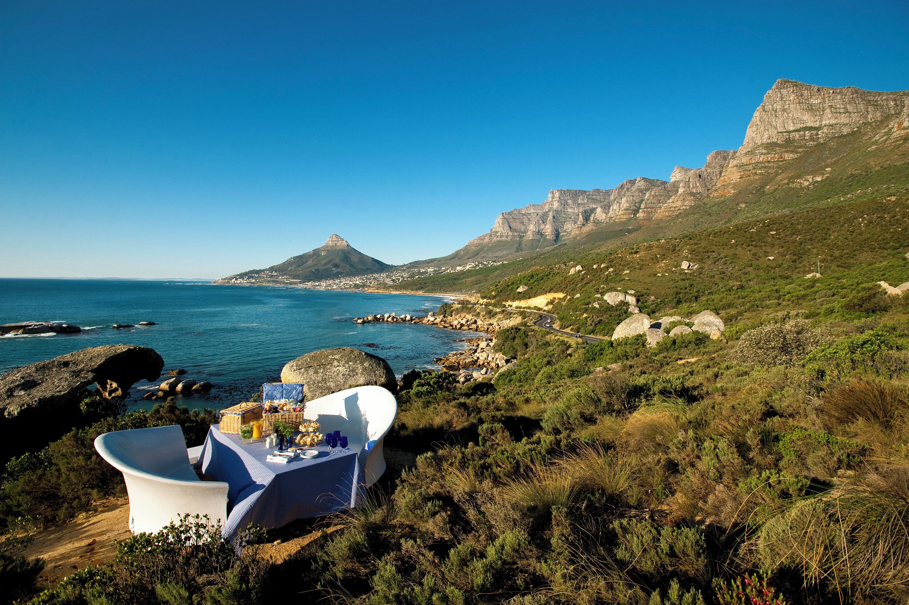A seaside dining setup with a table and chairs, overlooking a coastal landscape with cliffs and a clear sky.