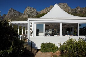 A white pavilion-style building with large windows and a balcony, surrounded by greenery and mountains in the background.