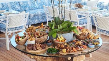Una mesa redonda de vidrio con diversos platos de comida a la parrilla y acompañamientos, rodeada de sillas de mimbre blanco y un sofá azul con estampado en un espacio exterior luminoso y soleado.
