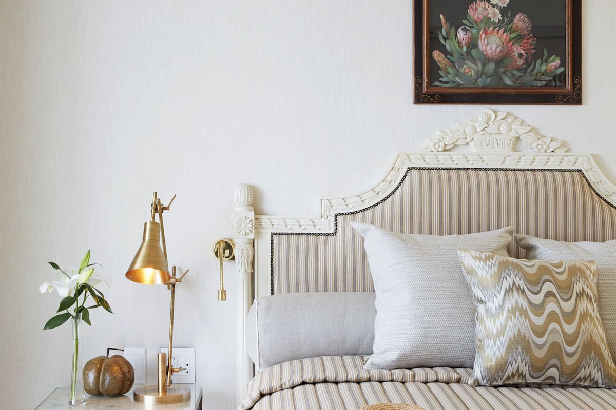 A neatly arranged bedroom with a striped bedspread, a bedside lamp, a vase with flowers, and a woven hat on the bed.