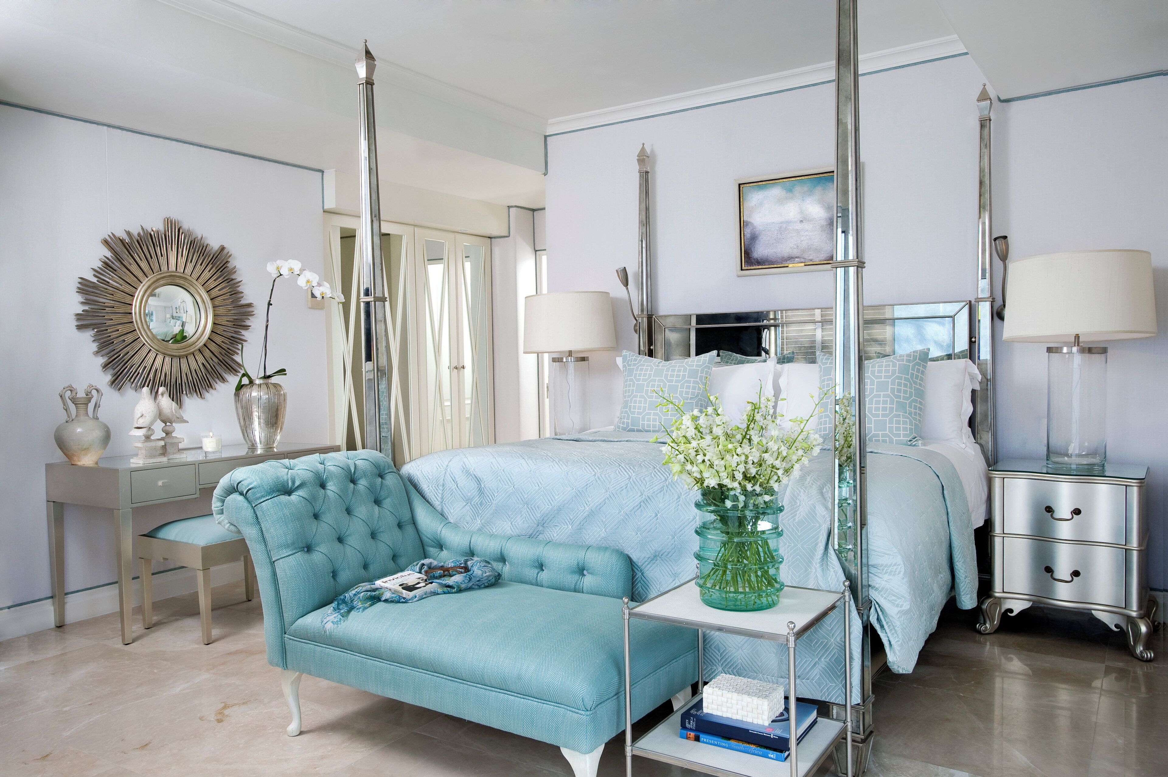 A bedroom with a bed, a blue sofa, a bedside table, a nightstand, a mirror, and a vase with flowers.