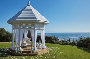 A gazebo with a white lattice roof and a wooden platform, set on a grassy area with white flowers, overlooking the ocean.