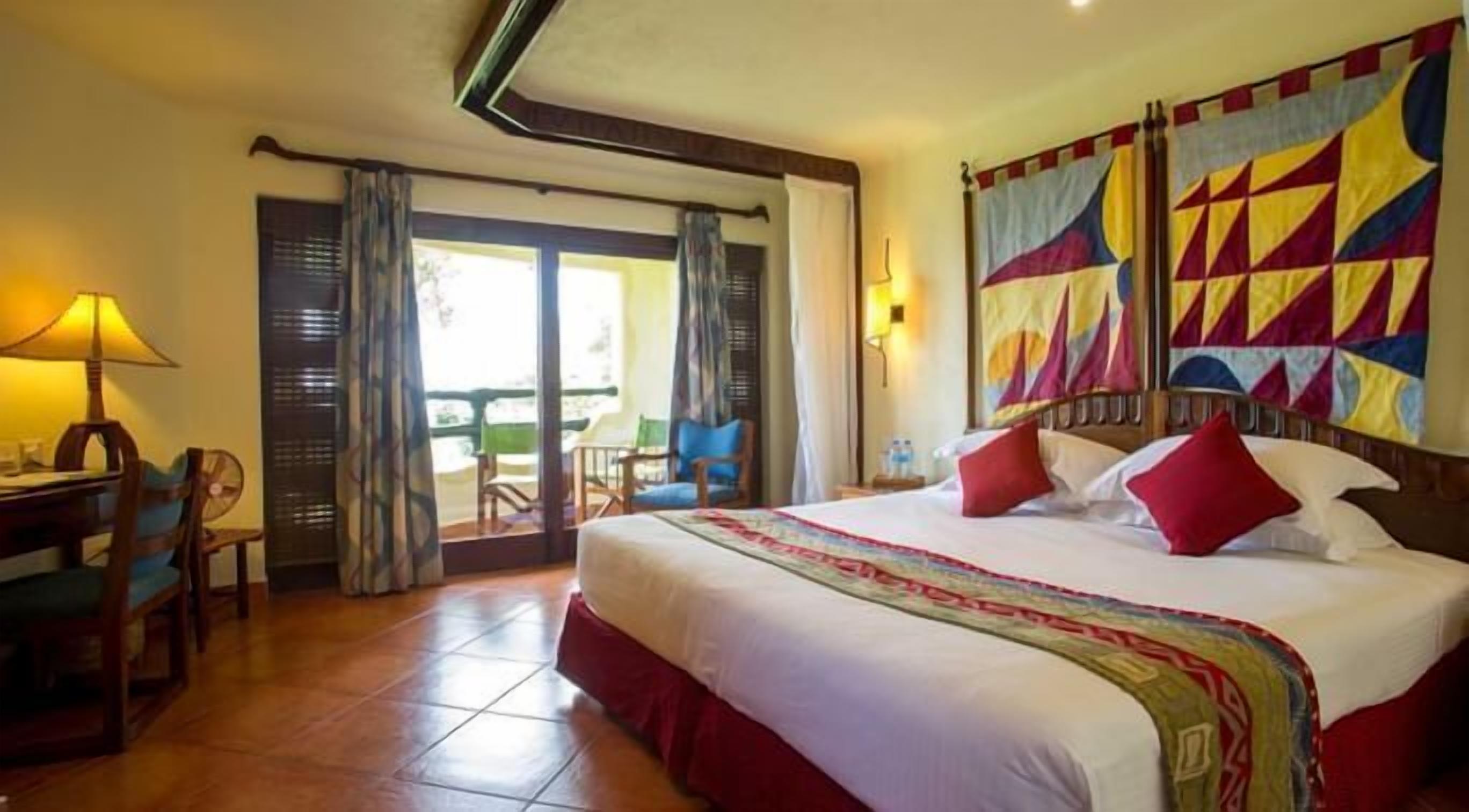 A hotel room with a large bed, a desk with a lamp, and a view of the outdoors through a sliding door.