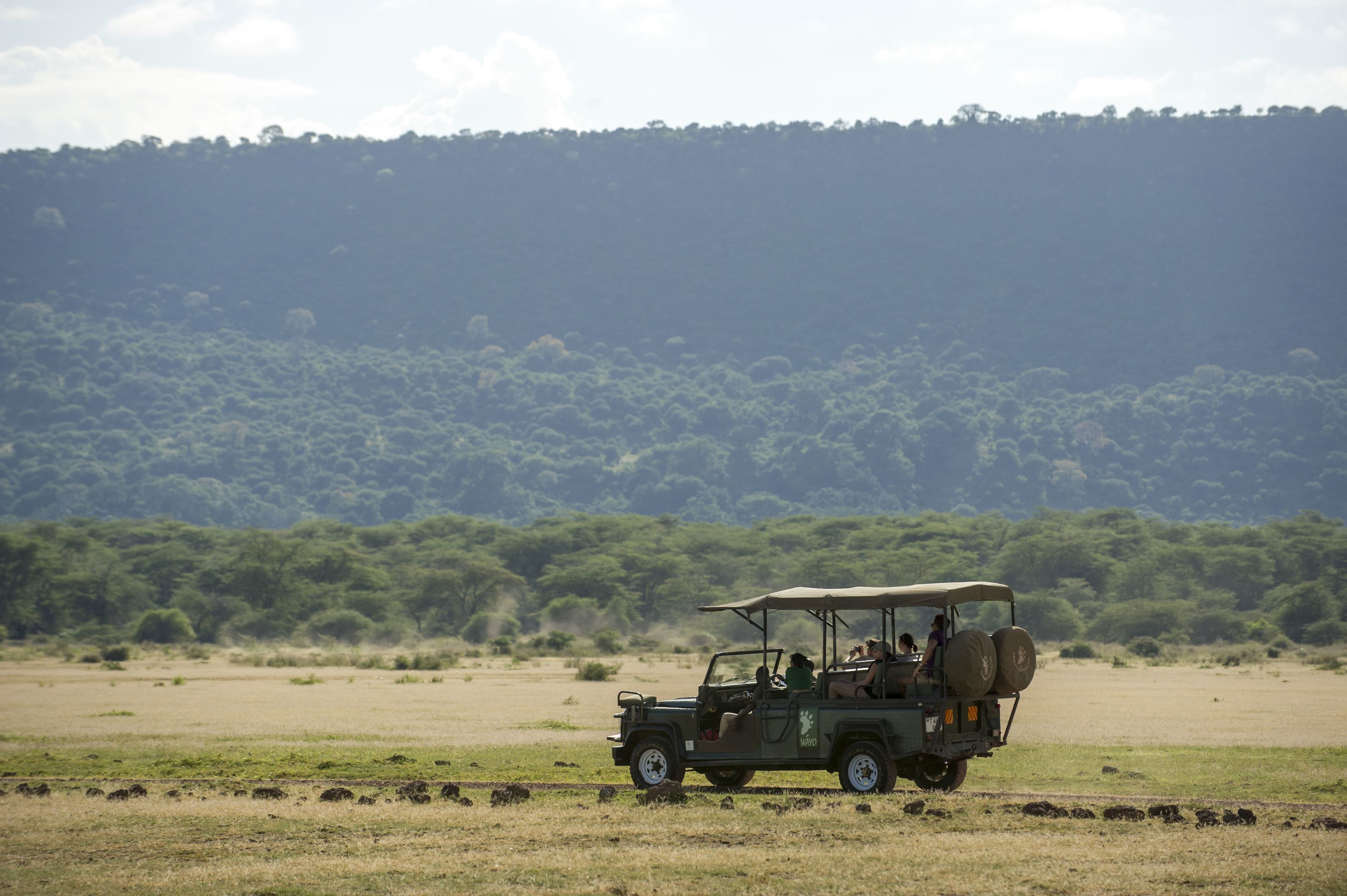 A safari vehicle with passengers, driving on a dirt road with a forested hill in the background.