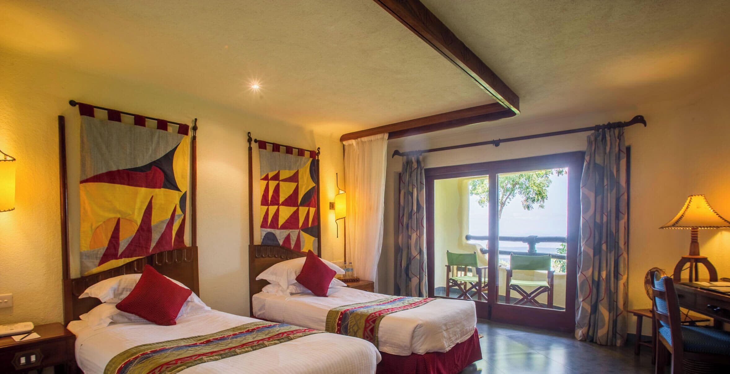 A hotel room with two beds, a desk, a chair, a lamp, and a view of the outdoors.