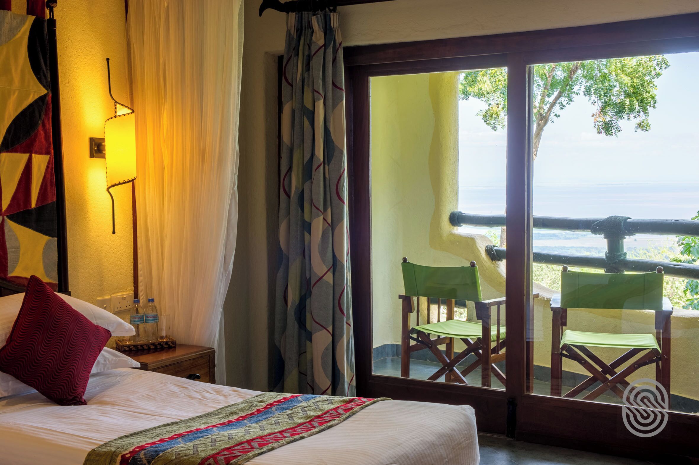 Habitación de hotel con cama, mesita de noche, lámpara, ventana con vista al mar y balcón con sillas.