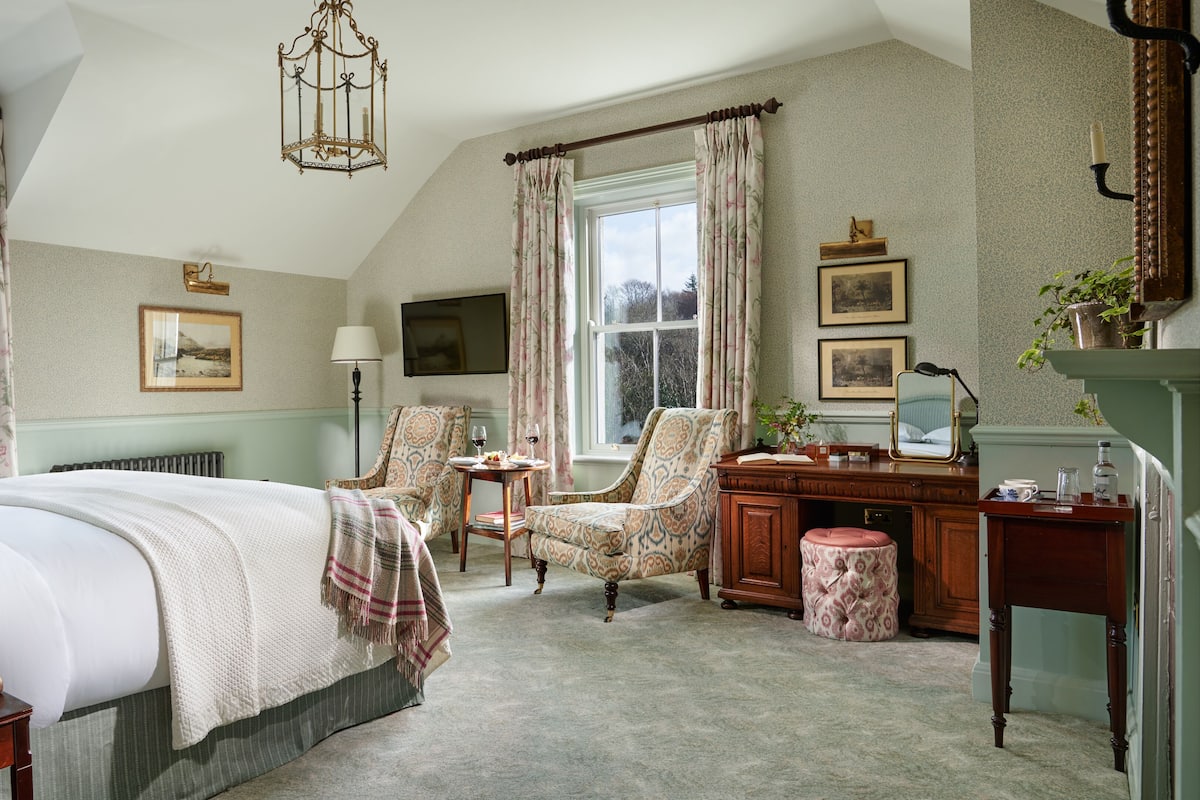 Un dormitorio elegante con una cama grande, un escritorio, una silla y un taburete pequeño. Hay una ventana con cortinas, una lámpara y un cuadro en la pared.