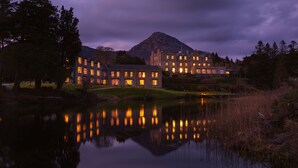 A large, multi-story building with illuminated windows, reflecting on a calm body of water, surrounded by trees and a mountain in the background. - Ballynahinch Castle Hotel (Ballynahinch)