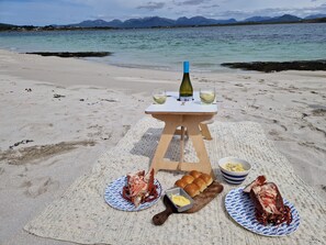 Una preparación para picnic en la playa, con una botella de vino, copas, pan, langosta y mantequilla sobre una manta.
