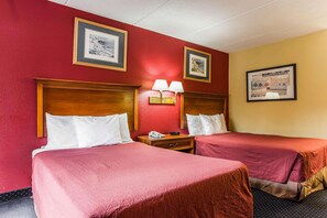 A hotel room with two beds, each with a wooden headboard and white pillows. There is a nightstand with a lamp between the beds. - Rodeway Inn Charlotte (Charlotte)