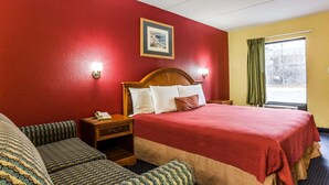 A hotel room with a large bed, a wooden headboard, a bedside table with a lamp, a nightstand, and a window with curtains.