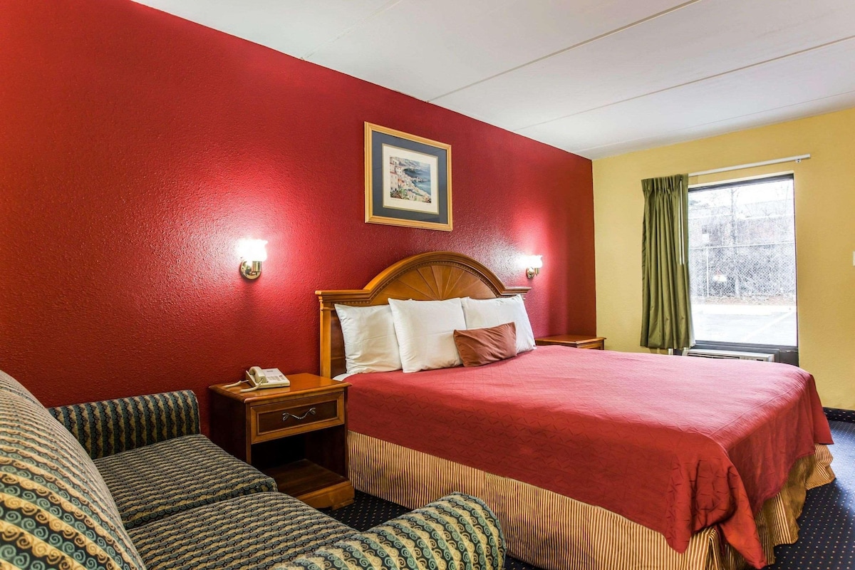 A hotel room with a large bed, a wooden headboard, a bedside table with a lamp, a nightstand, and a window with curtains.