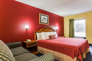 A hotel room with a large bed, a wooden headboard, a bedside table with a lamp, a nightstand, and a window with curtains. - Rodeway Inn Charlotte (Charlotte)