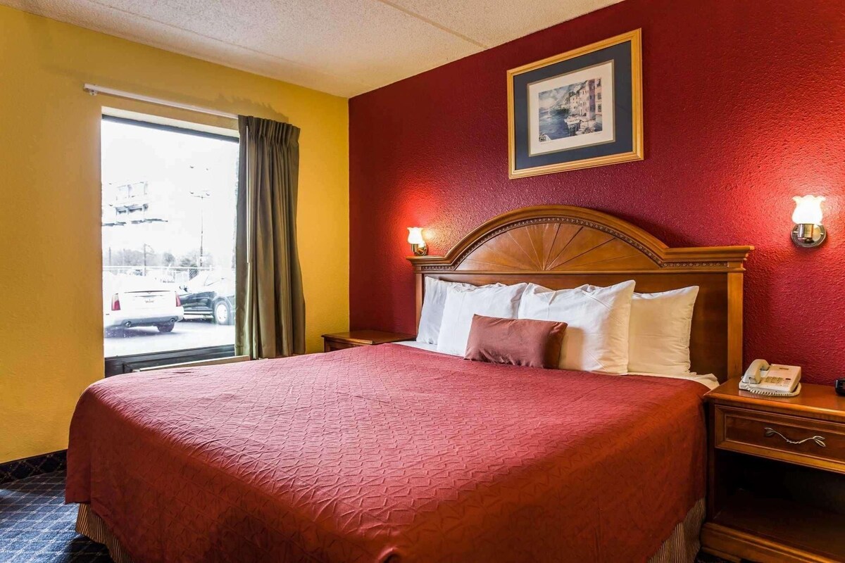 A hotel room with a large bed, a wooden headboard, a nightstand with a phone, and a window with a view of the street.