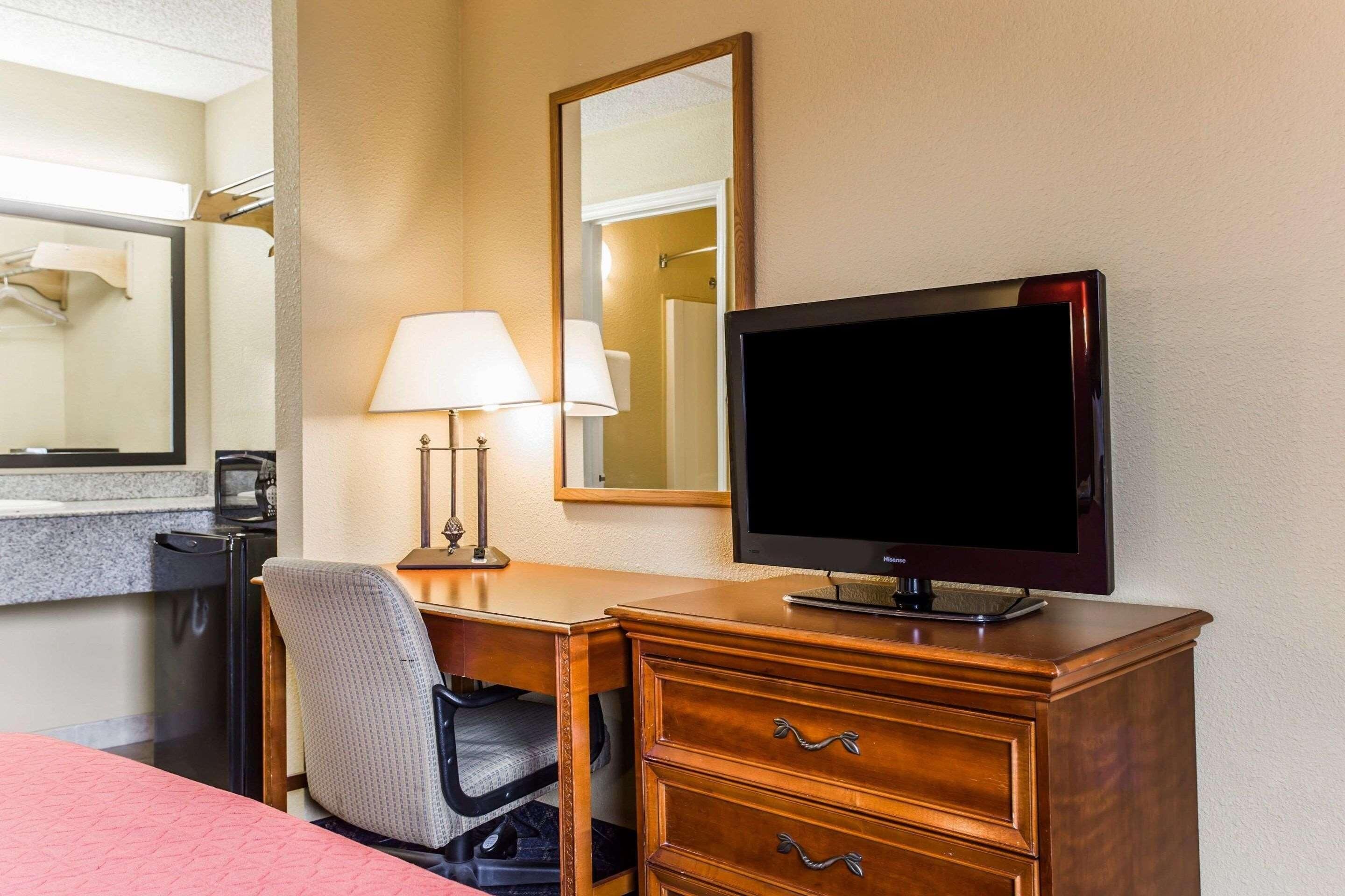 A hotel room with a wooden desk, a television, a lamp, a chair, and a bed with a pink bedspread. - Rodeway Inn Charlotte (Charlotte)