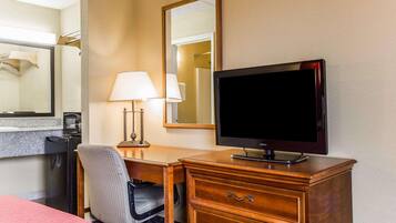 A hotel room with a wooden desk, a television, a lamp, a chair, and a bed with a pink bedspread.