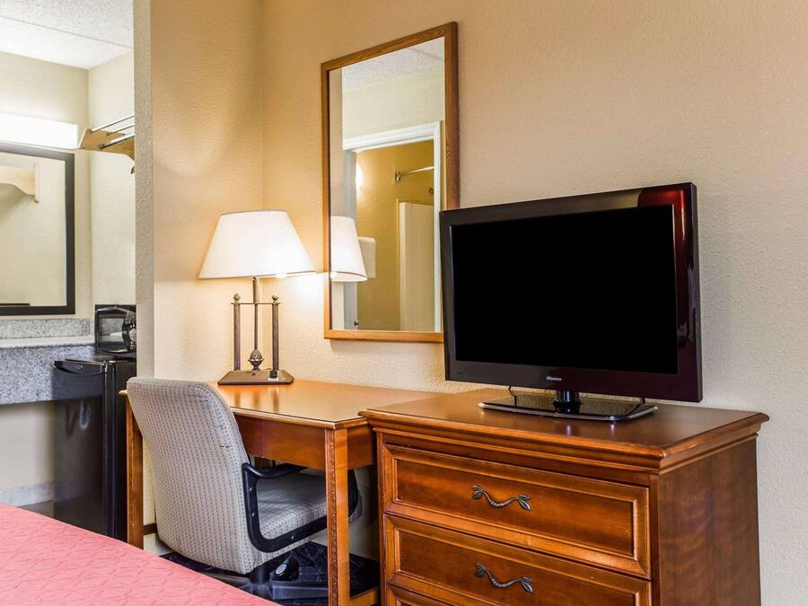 A hotel room with a wooden desk, a television, a lamp, a chair, and a bed with a pink bedspread.