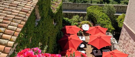 A terrace with red umbrellas and potted plants, overlooking a courtyard with a stone wall and greenery.