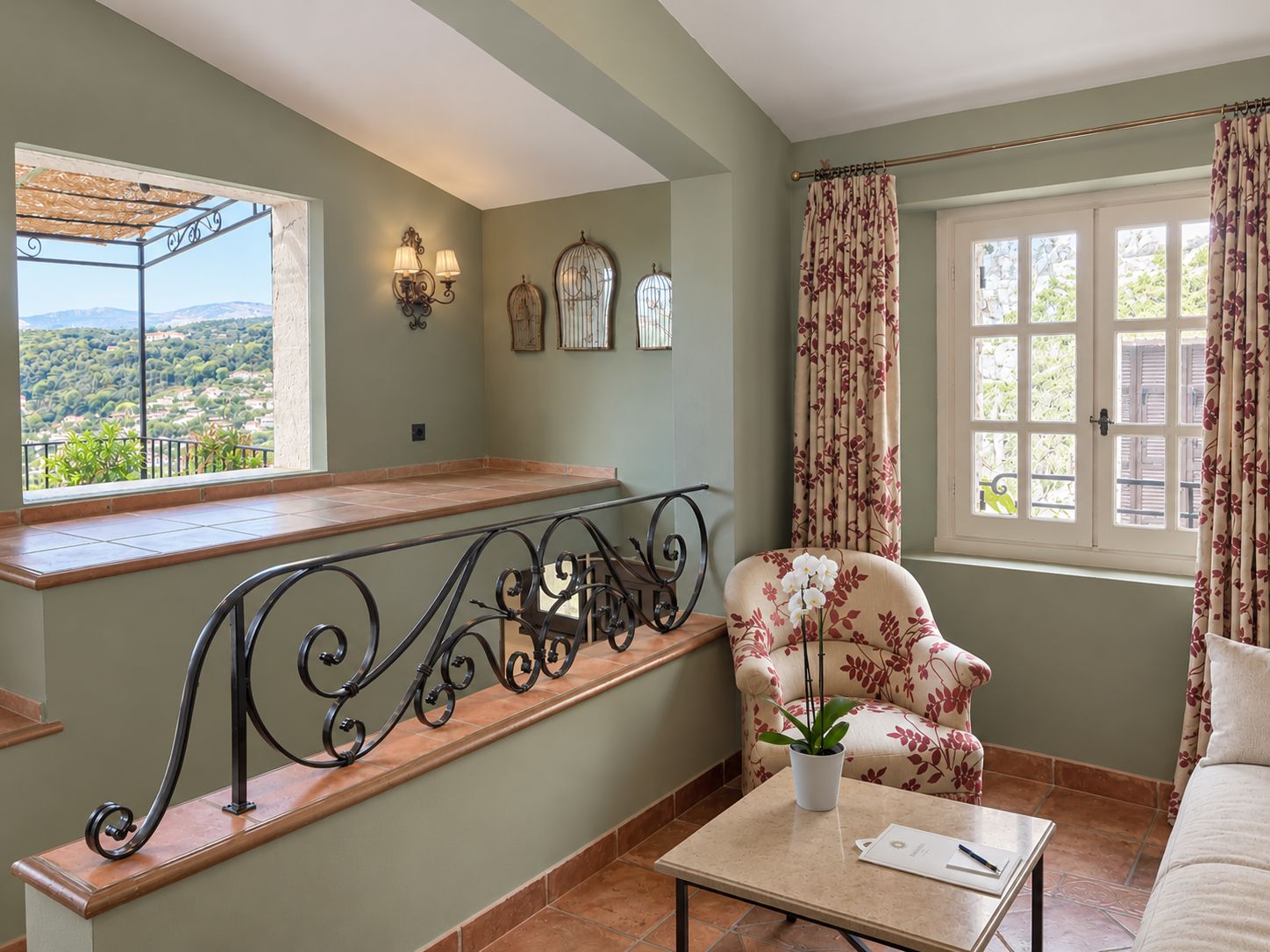 Une pièce avec un balcon, une chaise à motifs floraux, une table avec une plante et une fenêtre avec des rideaux.