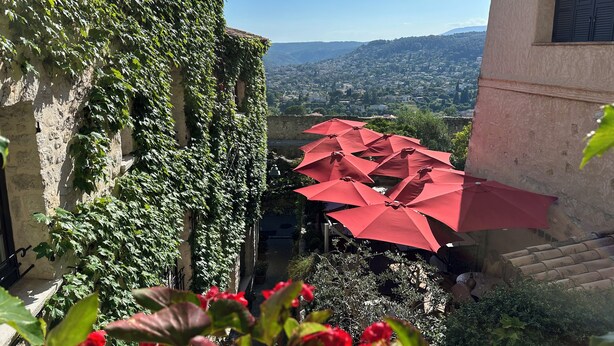 A courtyard with red umbrellas, a stone wall with greenery, and a view of a distant town. - Le Saint Paul (Saint-Paul-de-Vence)
