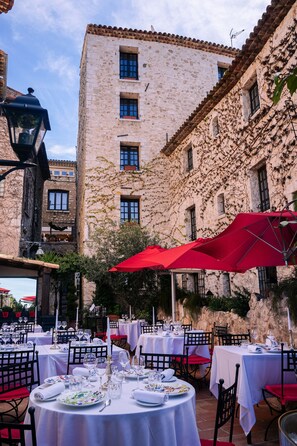 Un espace de restauration en plein air, avec des tables dressées pour un repas, entouré de bâtiments en pierre dotés de nombreuses fenêtres et de balcons.