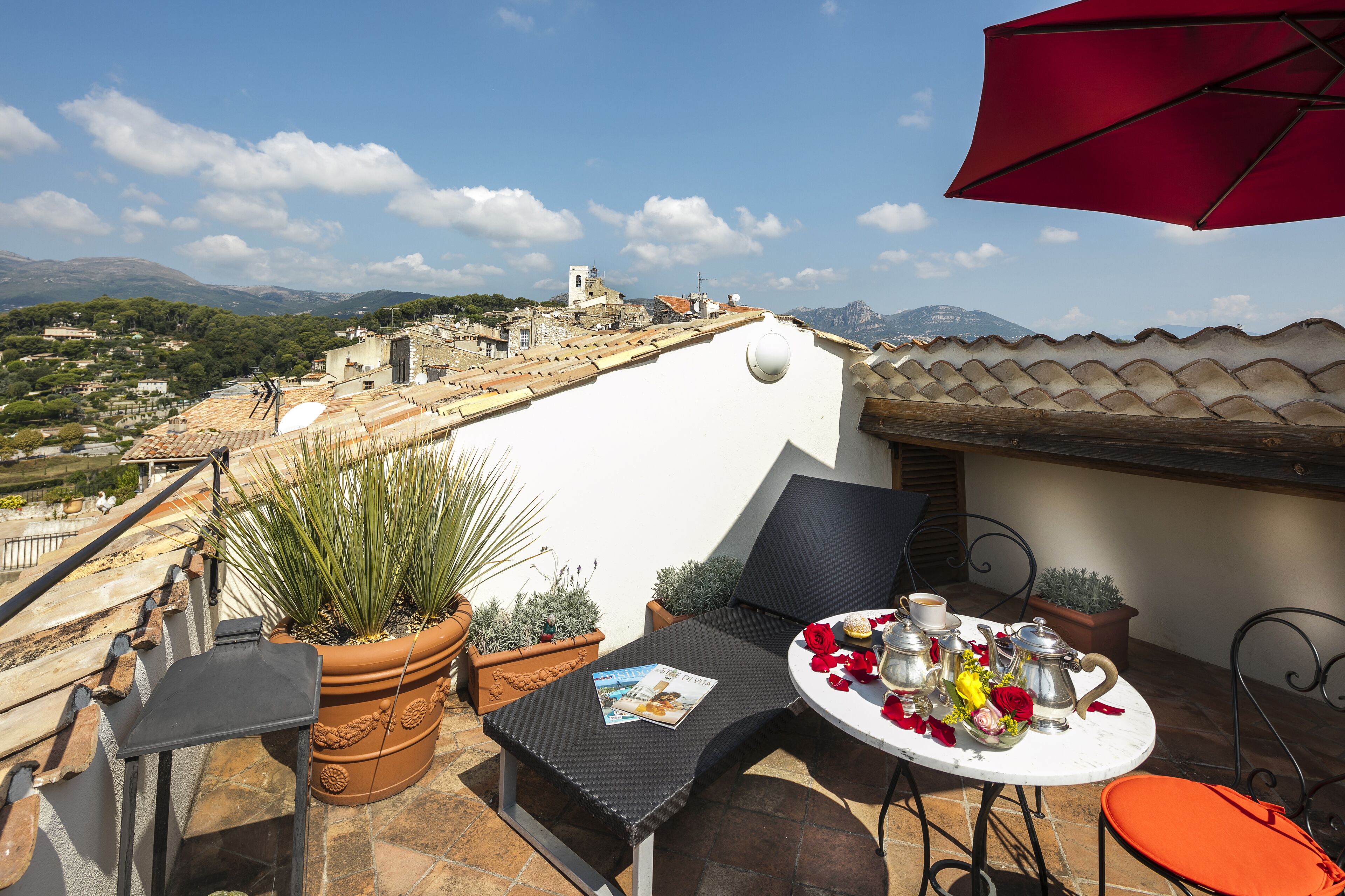 Um terraço com uma mesa posta para dois, plantas em vasos e vista para uma cidade e montanhas.