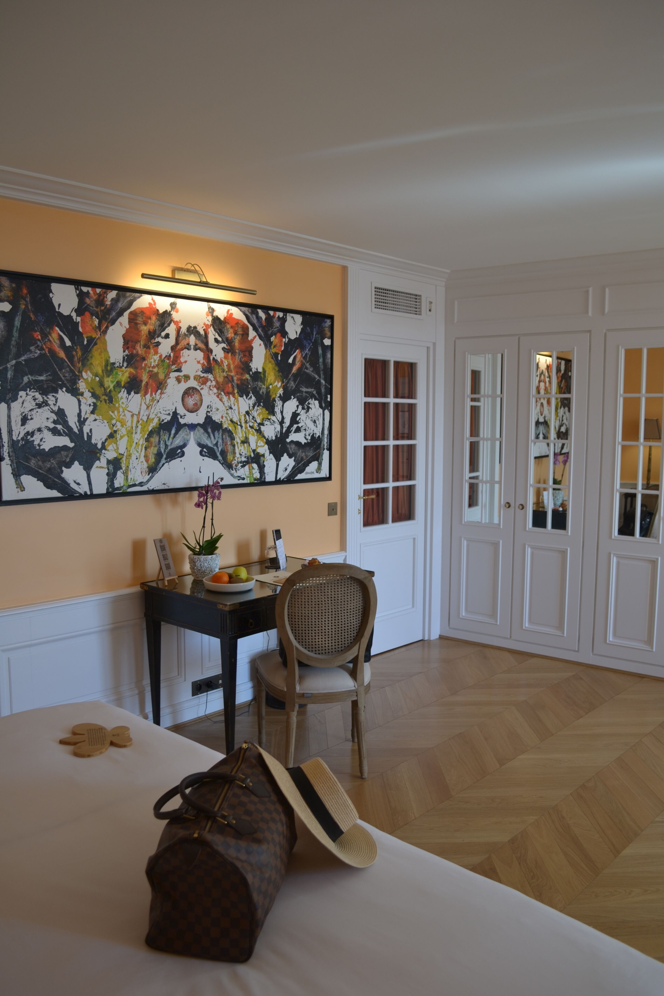 A hotel room with a bed, a desk with a chair, a large floral painting, and a wardrobe with glass doors.