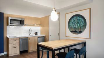 A modern kitchen with a microwave, dishwasher, wooden cabinets, a white countertop, and a dining area with a wooden table and blue chairs.