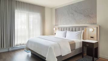 A hotel room with a large bed, a headboard with a wall art piece, a bedside table with a lamp, and a window with sheer curtains.