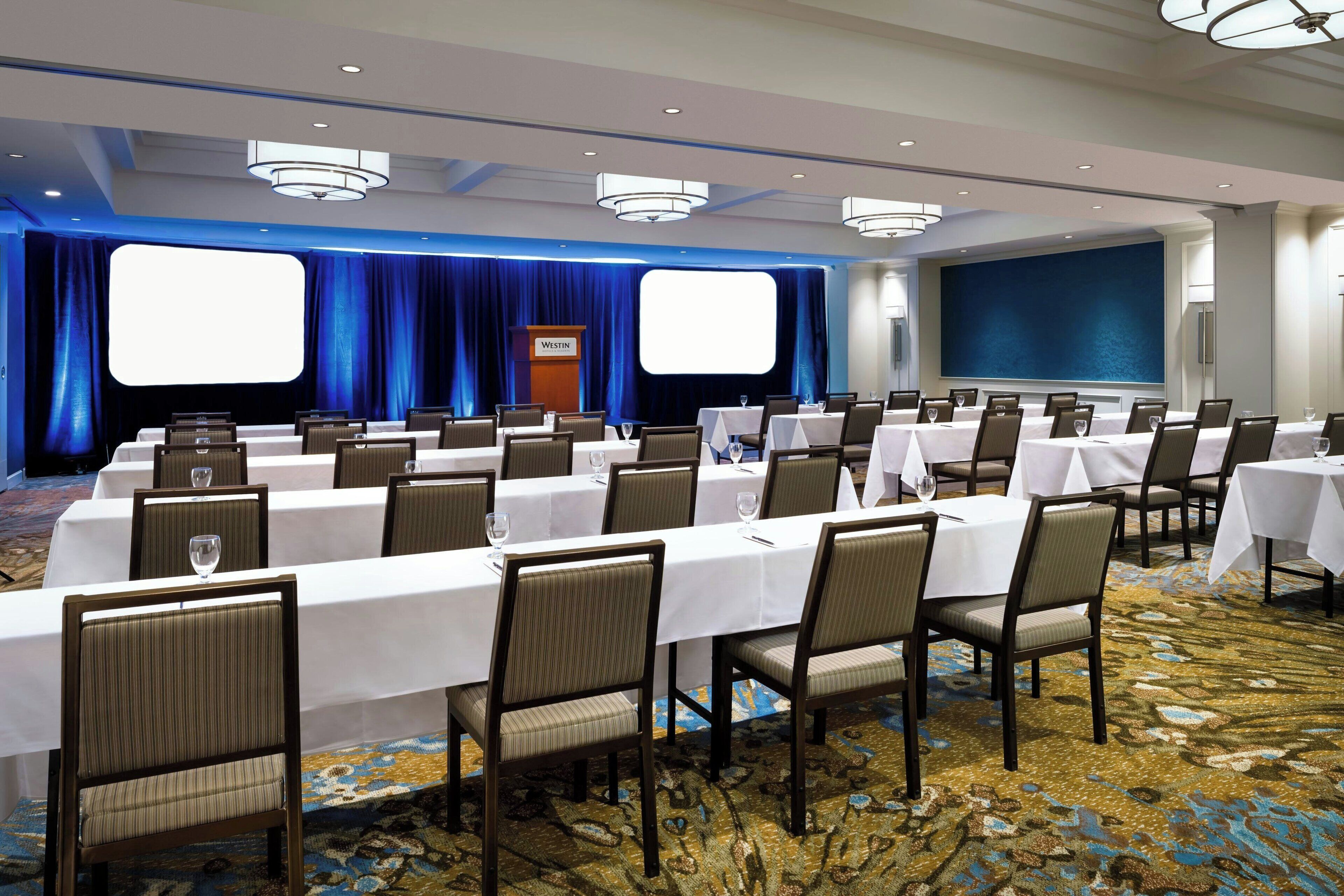 A conference room with multiple tables and chairs, a podium, and a large screen.