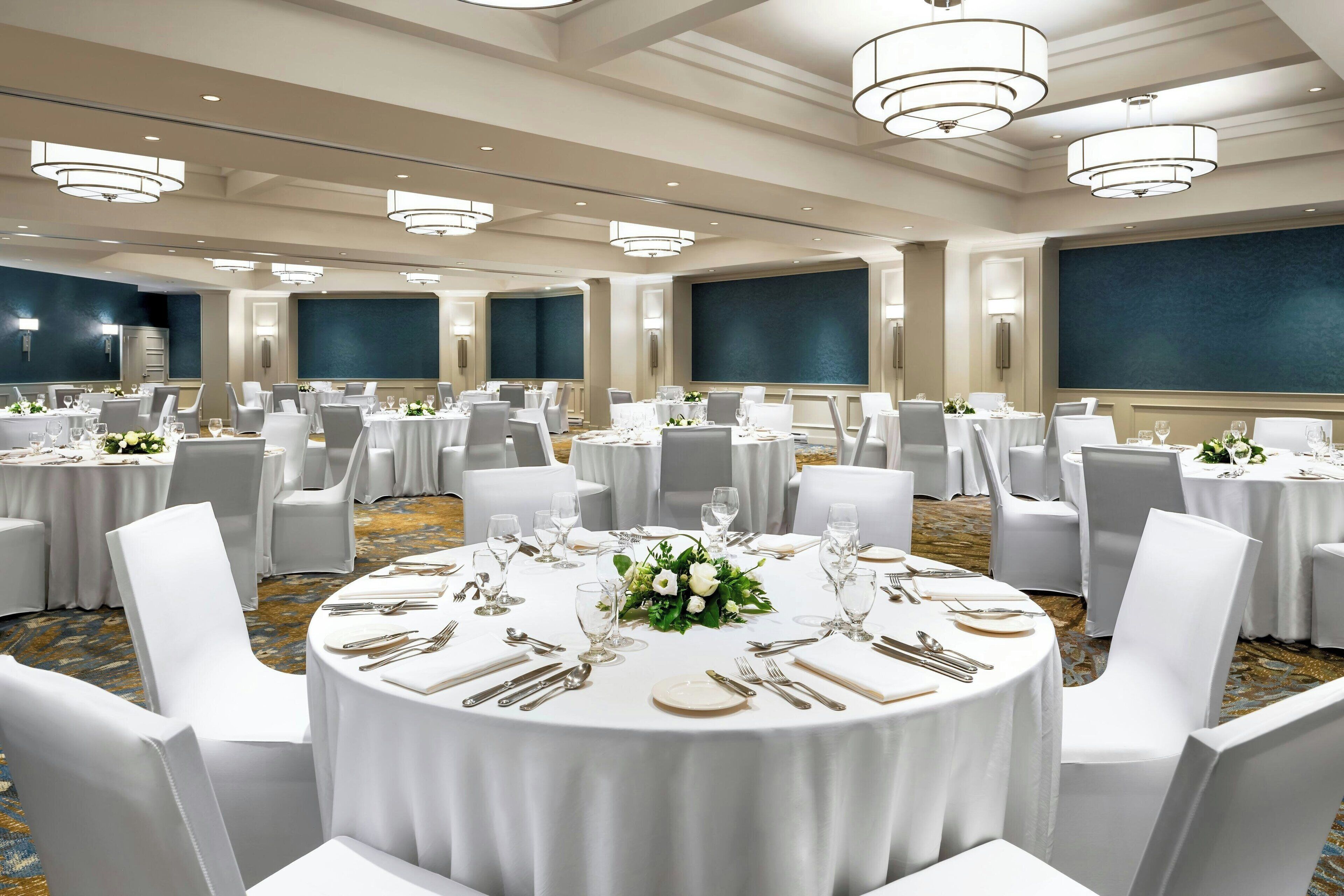 A banquet hall with round tables set for dining, white chairs, and chandeliers.