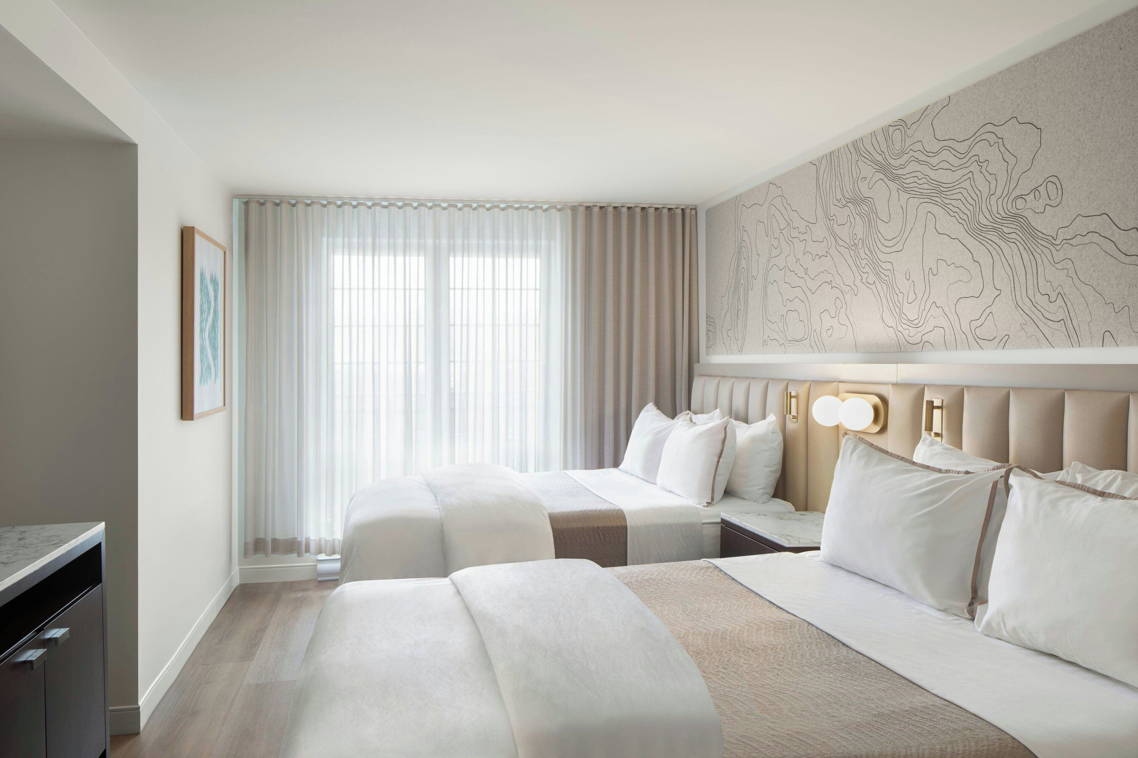 A hotel room with two beds, a large window with sheer curtains, a wall-mounted artwork, and a desk with a chair.