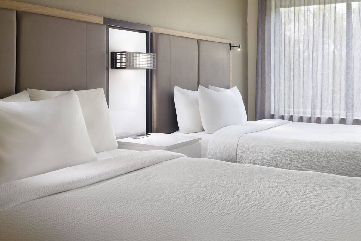 Habitación de hotel con dos camas, una mesita de noche y un amplio ventanal con cortinas traslúcidas.