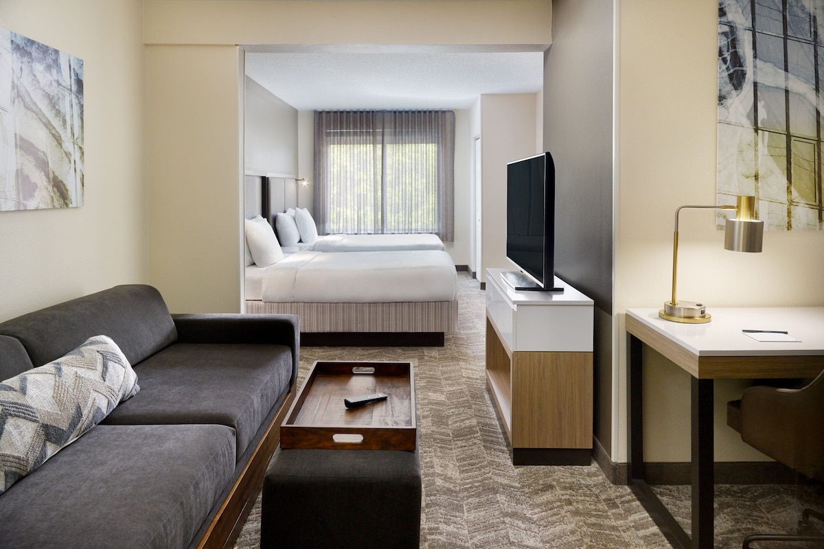 A hotel room with a sofa, a bed, a desk, a TV, and artwork on the walls.