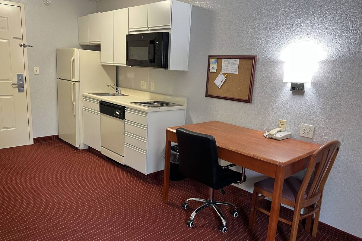 A compact kitchen with a microwave, sink, and dishwasher, a wooden dining table with chairs, and a corkboard with notes on the wall.