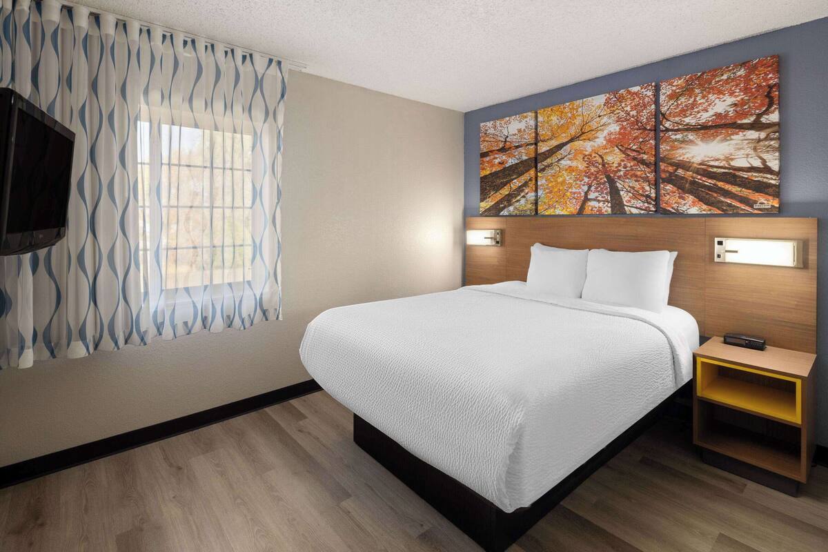 A hotel room with a bed, a TV mounted on the wall, a painting above the bed, and a nightstand with a lamp.