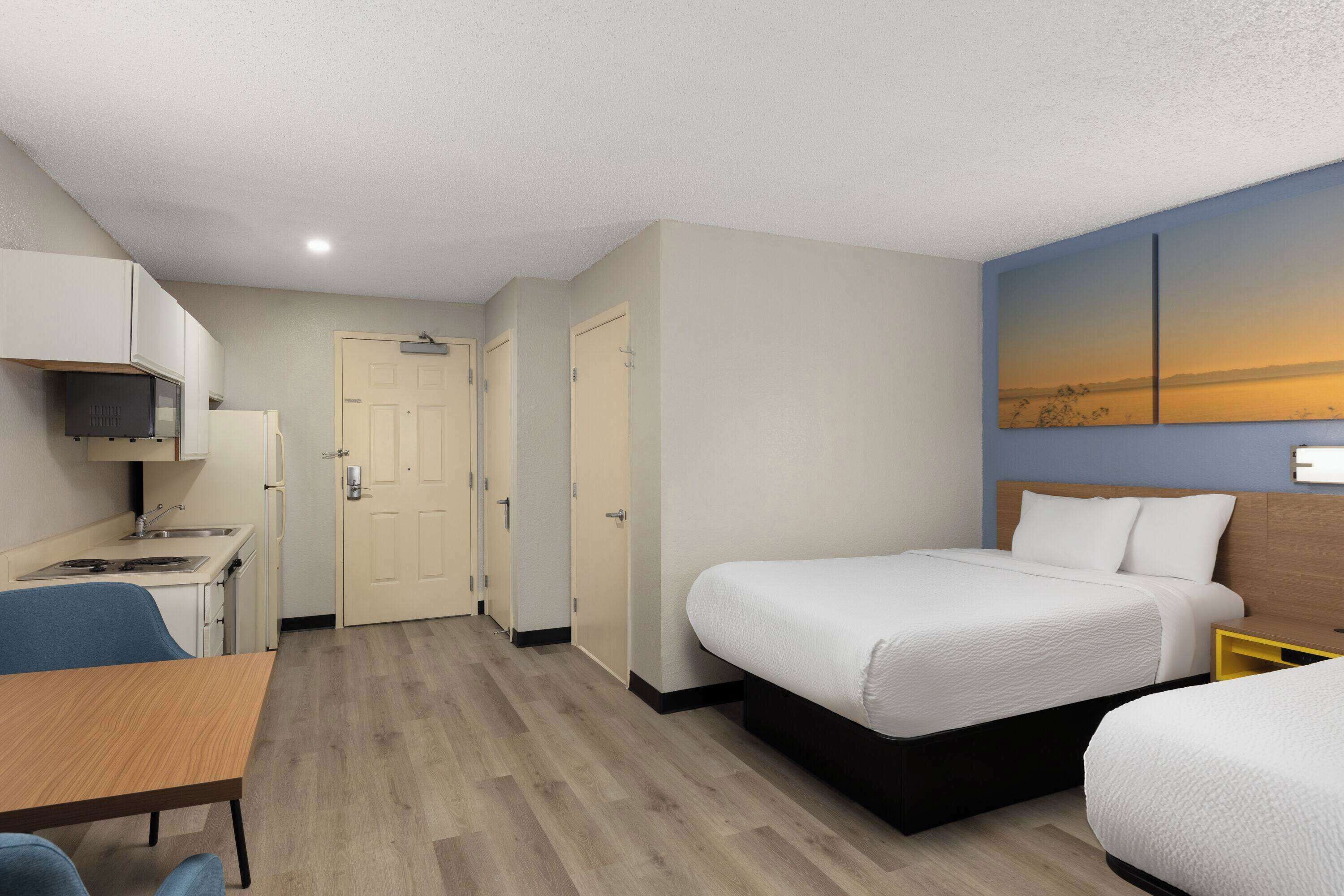 A hotel room with a bed, a desk, a chair, a small kitchenette, and a door leading to another room.