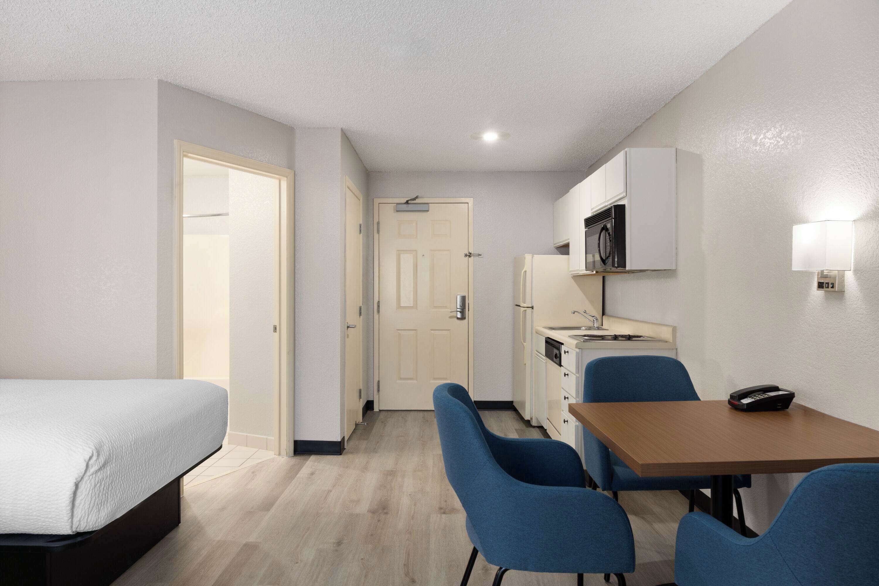 Habitación de hotel con cama, escritorio, dos sillas, microondas, refrigerador y puerta que da a otra habitación.
