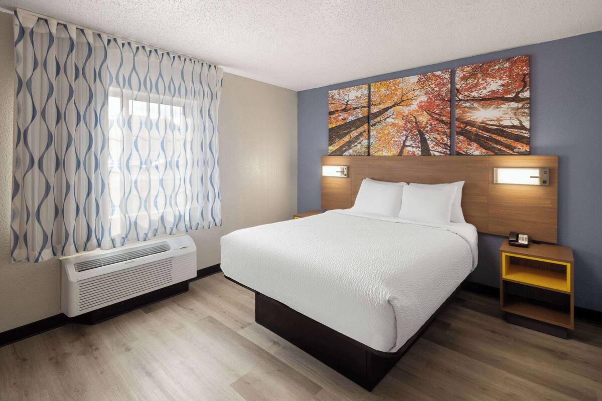 A hotel room with a bed, a bedside table with a phone, a wall-mounted light, and a window with patterned curtains.