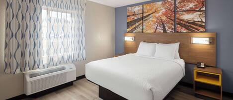 A hotel room with a bed, a bedside table with a phone, a wall-mounted light, and a window with patterned curtains.