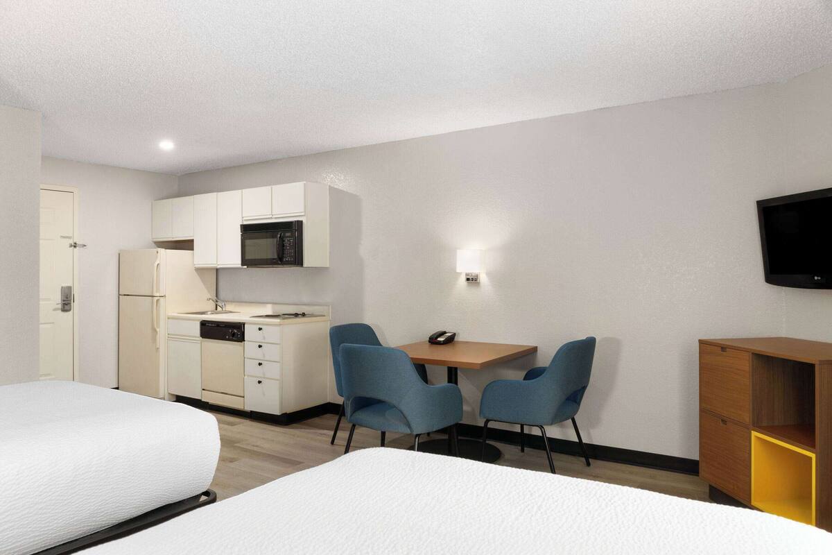 A hotel room with a bed, a desk with two chairs, a microwave, a refrigerator, and a TV mounted on the wall.