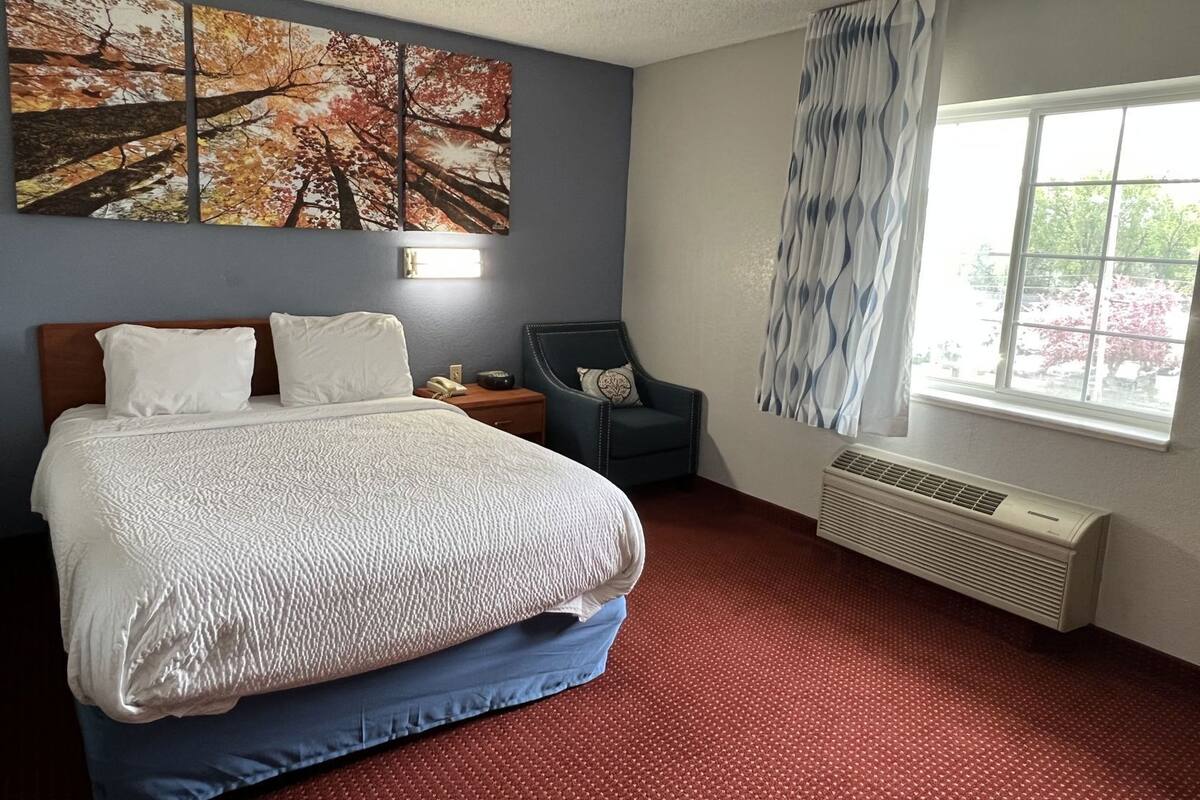 A hotel room with a bed, a chair, a window with curtains, a wall-mounted air conditioner, and a painting above the bed.